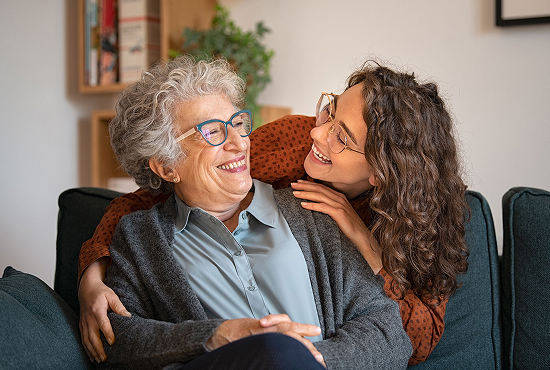 An elderly woman and a younger woman, both with curly hair and glasses, are sitting on a couch. They are smiling and embracing warmly. Bookshelves and a plant are in the blurred background, creating a cozy atmosphere.