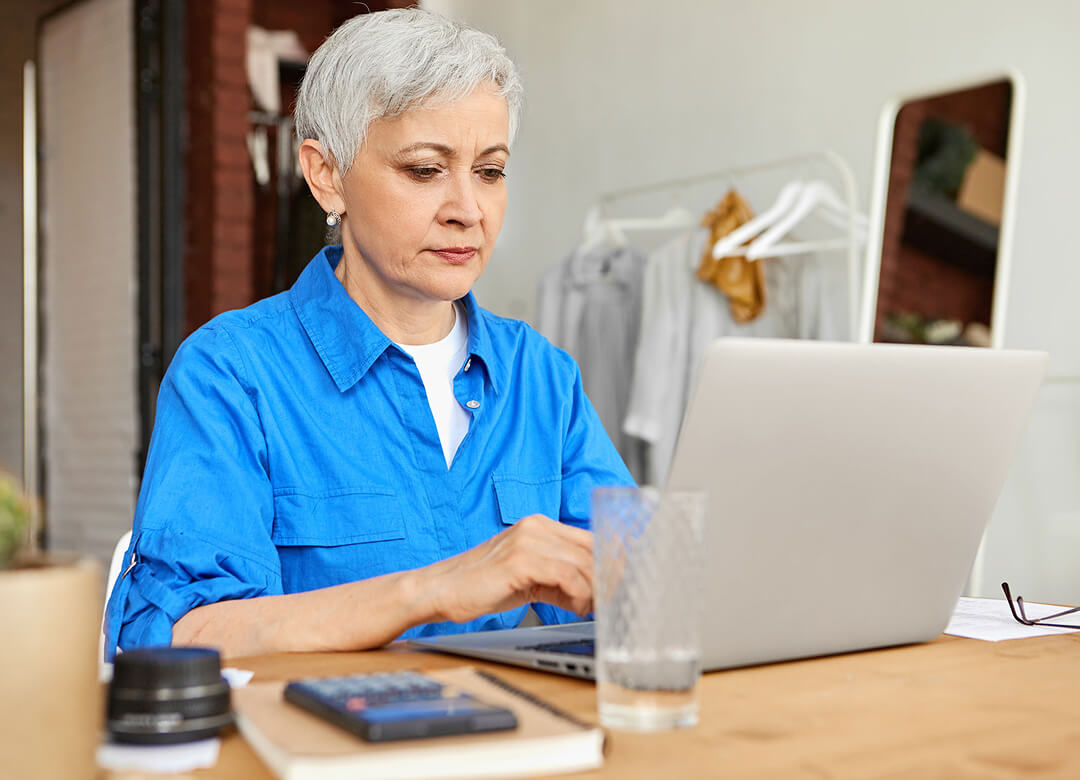 Person with short gray hair in a blue shirt working on a laptop at a table, with a glass of water, book, and camera lens nearby. Clothes are hanging in the background.