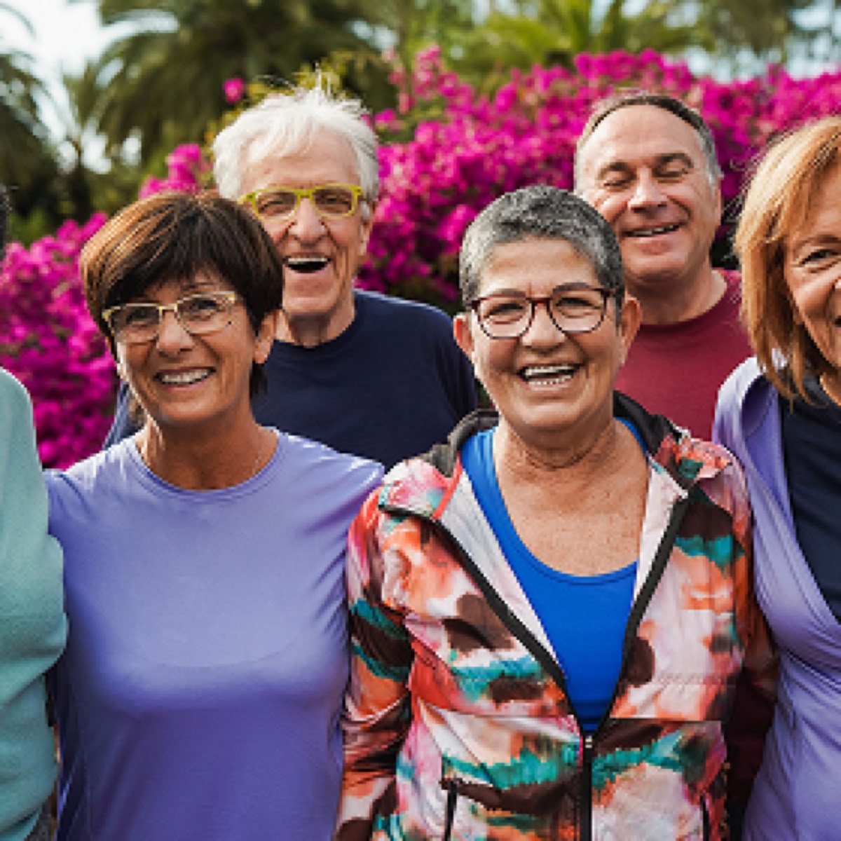 A group of six smiling adults stands together outdoors in front of vibrant pink flowers, wearing casual clothing and glasses.