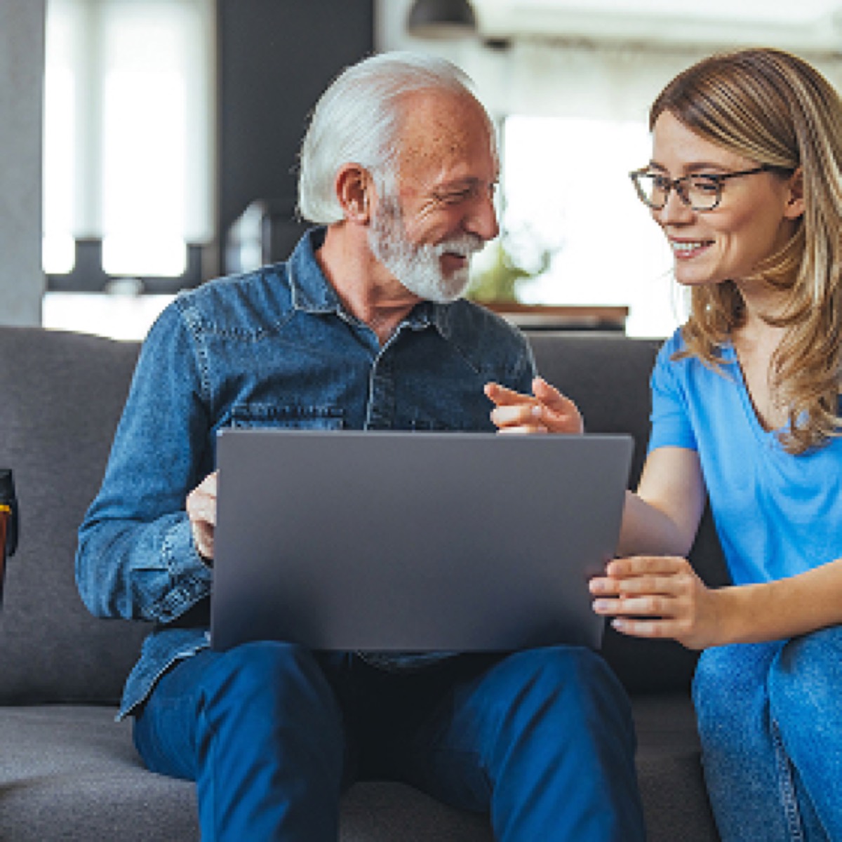 An elderly man and a young woman sit on a sofa, smiling while looking at a laptop.